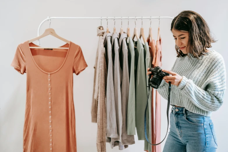 a woman standing in front of a rack of clothes, trending on pexels, holding a camera, colourised, wearing a light shirt, dlsr photo