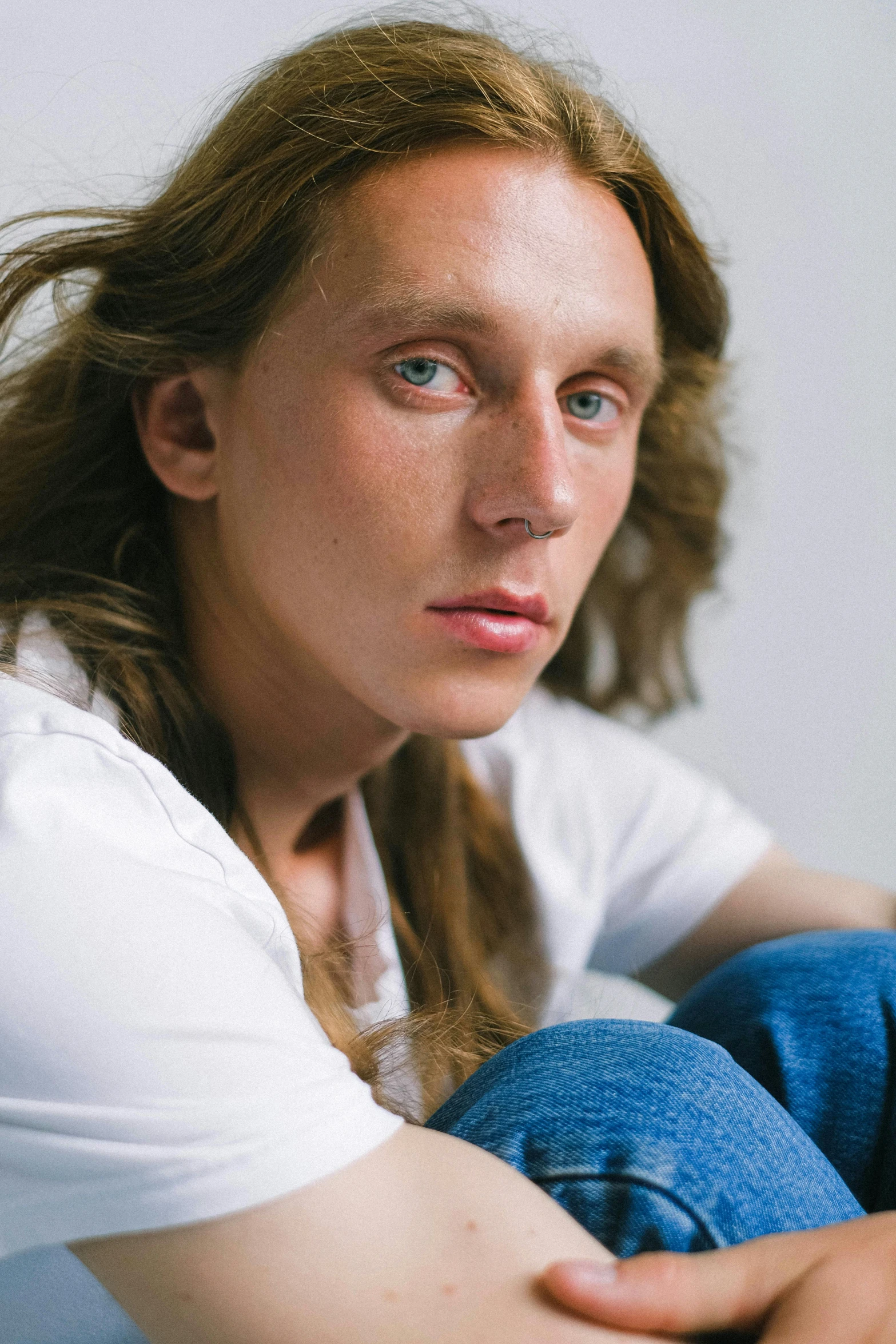 a close up of a person sitting on a bed, an album cover, inspired by Caspar van Wittel, white shirt and blue jeans, androgynous face, long mane, ryan mcginley