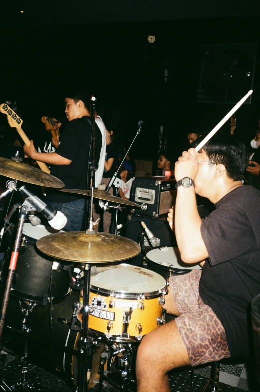 a man sitting on a stool in front of a drum set, an album cover, by Basuki Abdullah, unsplash, happening, punk concert, grainy low quality, a group of people, south jakarta