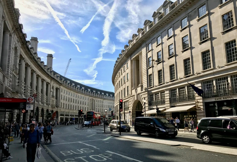 a group of people walking down a street next to tall buildings, by Rachel Reckitt, pexels contest winner, neoclassicism, portcullis, blue skies, square, lots of shops