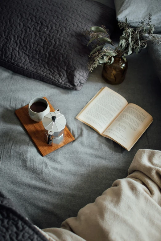 an open book sitting on top of a bed next to a cup of coffee, grey, slate, un made bed, dwell
