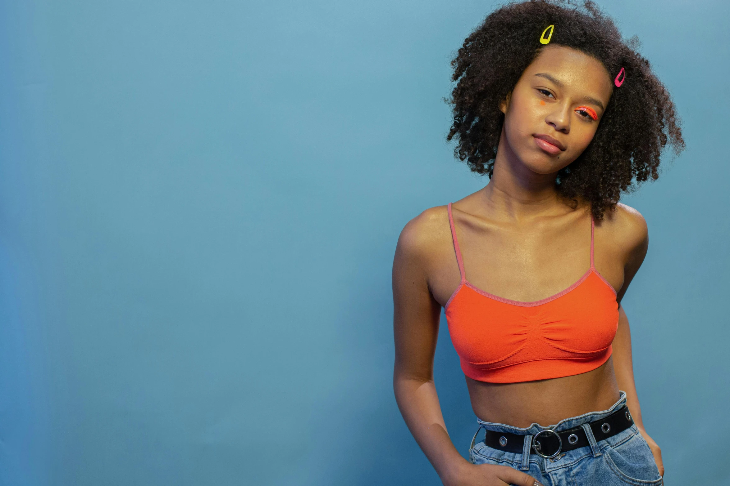 a beautiful young woman standing in front of a blue wall, inspired by Elsa Bleda, trending on pexels, renaissance, red bra, black teenage girl, orange neon, wearing yellow croptop