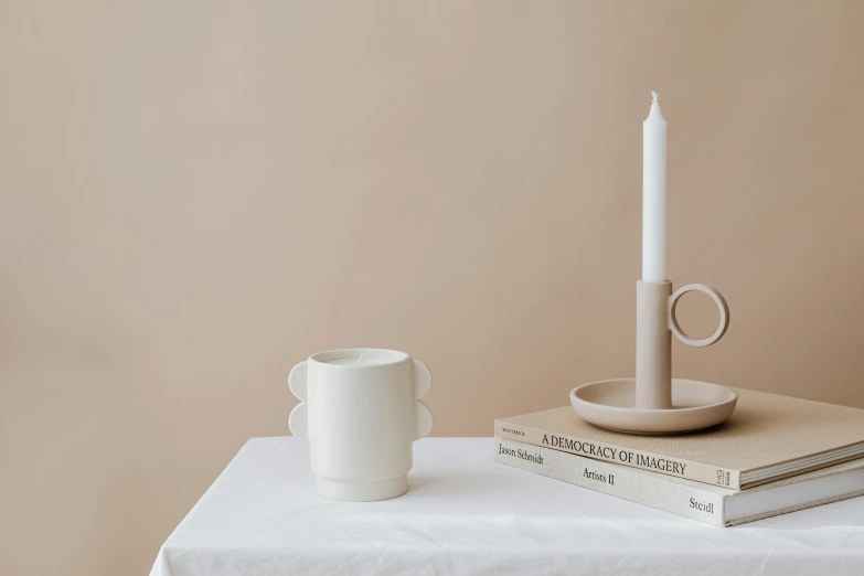 a white candle sitting on top of a table next to a stack of books, an abstract sculpture, inspired by Hendrik Gerritsz Pot, minimalism, with a white mug, holding a candle holder, pale beige sky, place setting