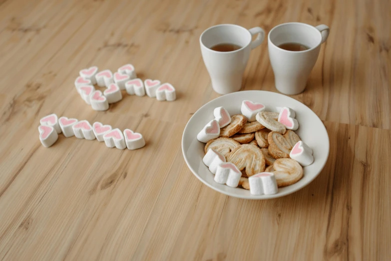 a white plate topped with marshmallows next to two cups of coffee, by Emma Andijewska, pexels contest winner, cookies, several hearts, alphabet soup, wood cups