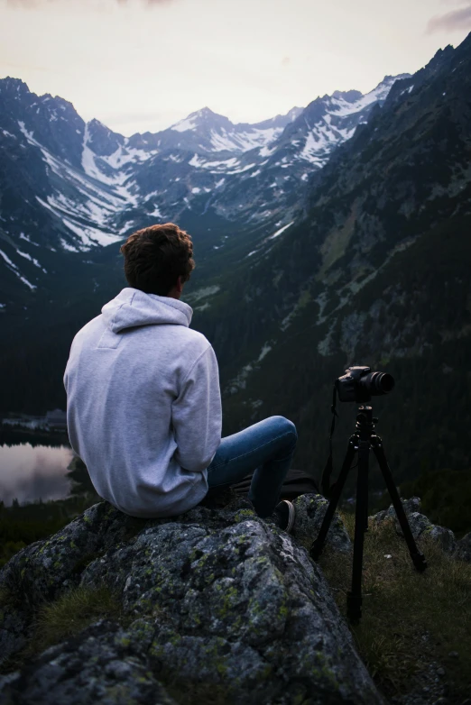 a man sitting on top of a rock next to a camera, inspired by Michael Komarck, unsplash contest winner, visual art, mountainous setting, sittin, looking at camera, man sitting facing away