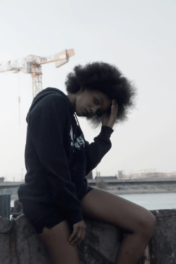 a woman sitting on a wall next to a body of water, an album cover, by Afewerk Tekle, trending on unsplash, black hoodie, big afro, intimidating pose, construction