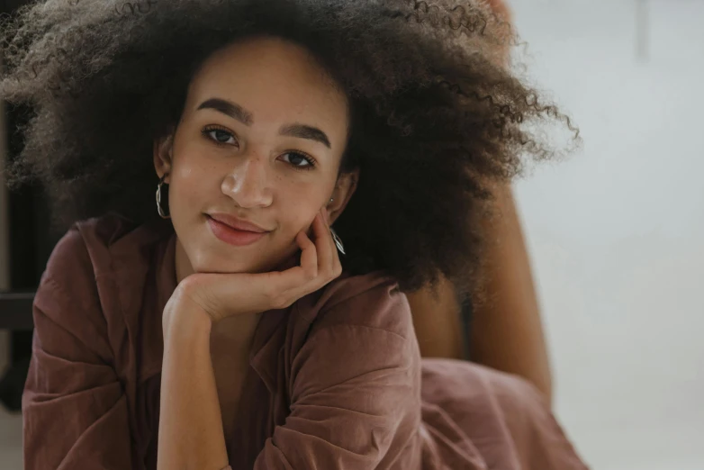 a beautiful young woman laying on top of a bed, trending on pexels, happening, long afro hair, rounded face, zoomed in, hand on her chin