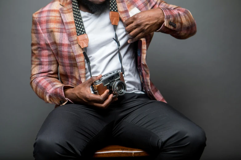 a man sitting on a stool holding a camera, trending on pexels, intricate leather suspenders, man is with black skin, wearing plaid shirt, subject detail: wearing a suit