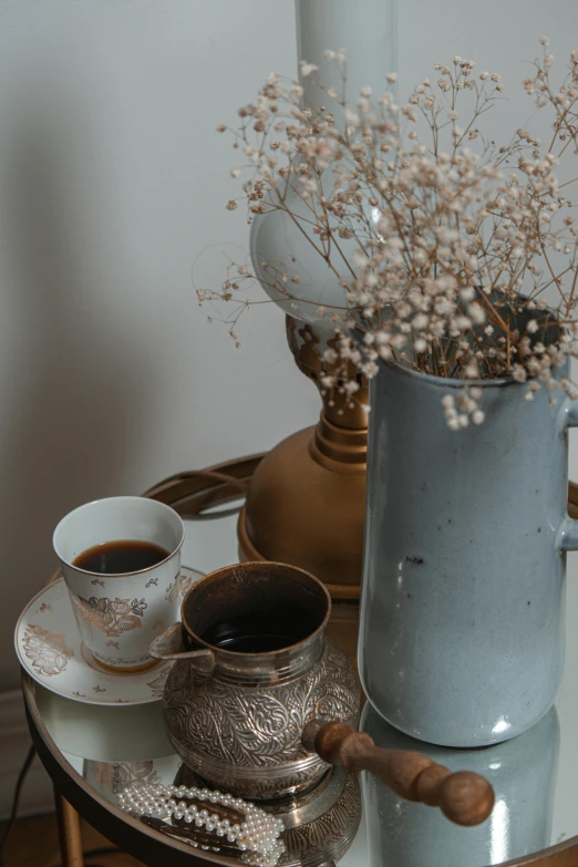 a coffee pot sitting on top of a table next to a cup of coffee, a still life, by Anna Haifisch, trending on unsplash, art nouveau, dried flowers, light grey blue and golden, middle eastern details, two cups of coffee