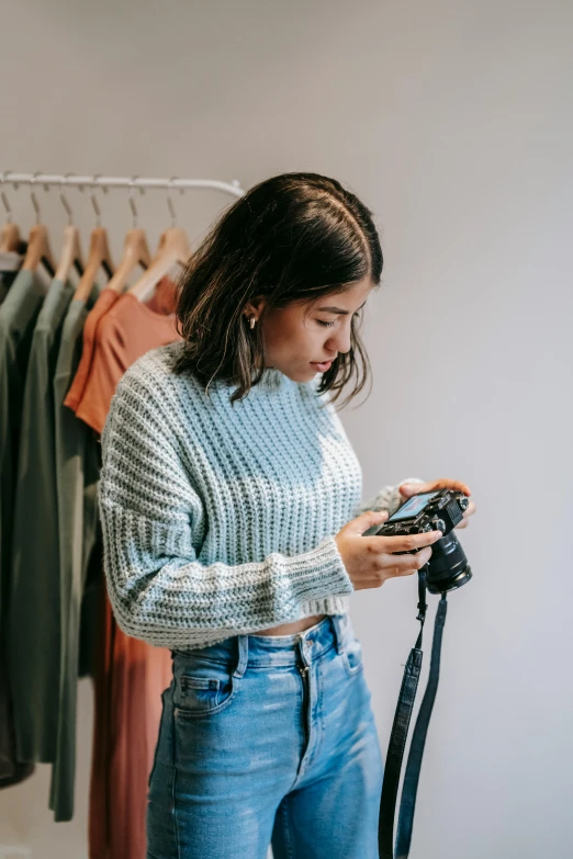 a woman standing in front of a rack of clothes holding a camera, wearing casual sweater, croptop, 2019 trending photo, focused photo