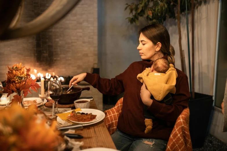 a woman sitting at a table with a baby in her arms, by Julia Pishtar, pexels contest winner, renaissance, candlelit restaurant table, cooking oil, autumn season, celebrating a birthday