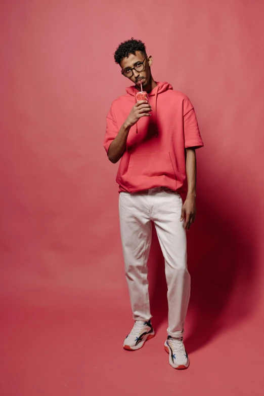 a man standing in front of a pink background, inspired by Louis Hersent, trending on pexels, white pants, red hoodie, man with glasses, holding a bottle