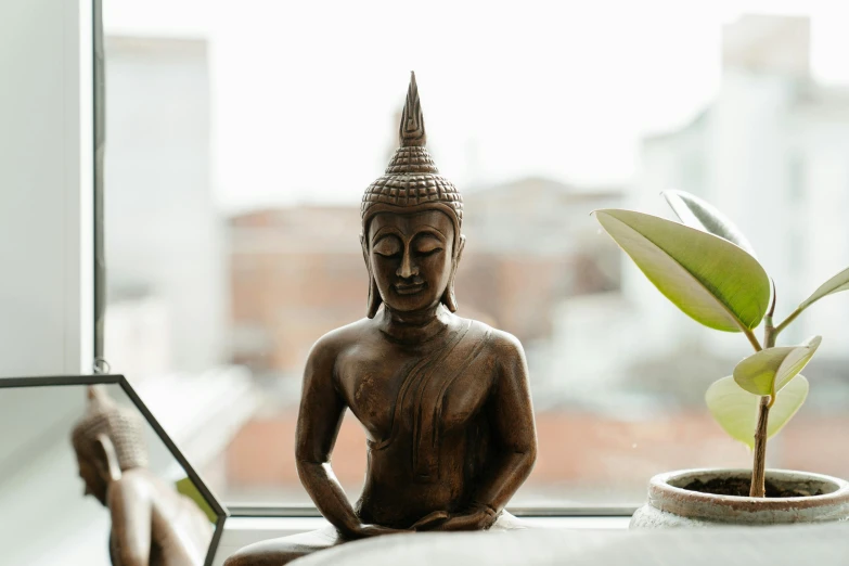 a buddha statue sitting next to a potted plant, a statue, unsplash, large open windows, brown, brightly-lit, windowsill