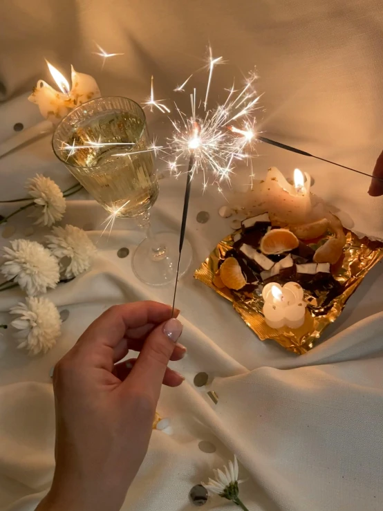 a close up of a person holding a sparkler, on a sumptuous tablecloth, full product shot, cosy vibes, charcoal and champagne