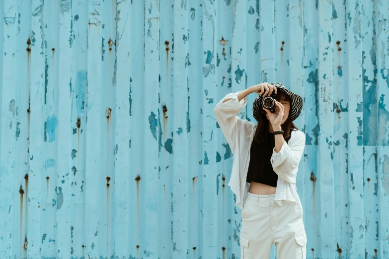 a woman taking a picture with a camera, pexels contest winner, minimalism, white and teal metallic accents, blue wall, caracter with brown hat, outdoor photo