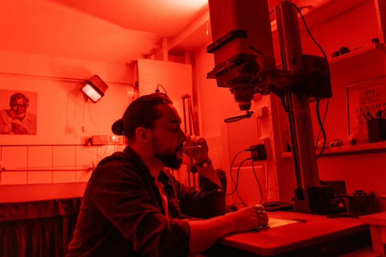 a man sitting at a table talking on a cell phone, by Breyten Breytenbach, pexels contest winner, holography, in an underground laboratory, red light, microscope, student