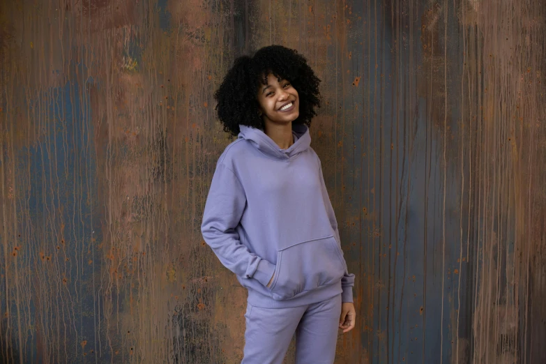 a woman standing in front of a metal wall, inspired by Itō Seiu, trending on pexels, happening, wearing a purple sweatsuit, wearing a grey hooded sweatshirt, muted pastels, looking happy