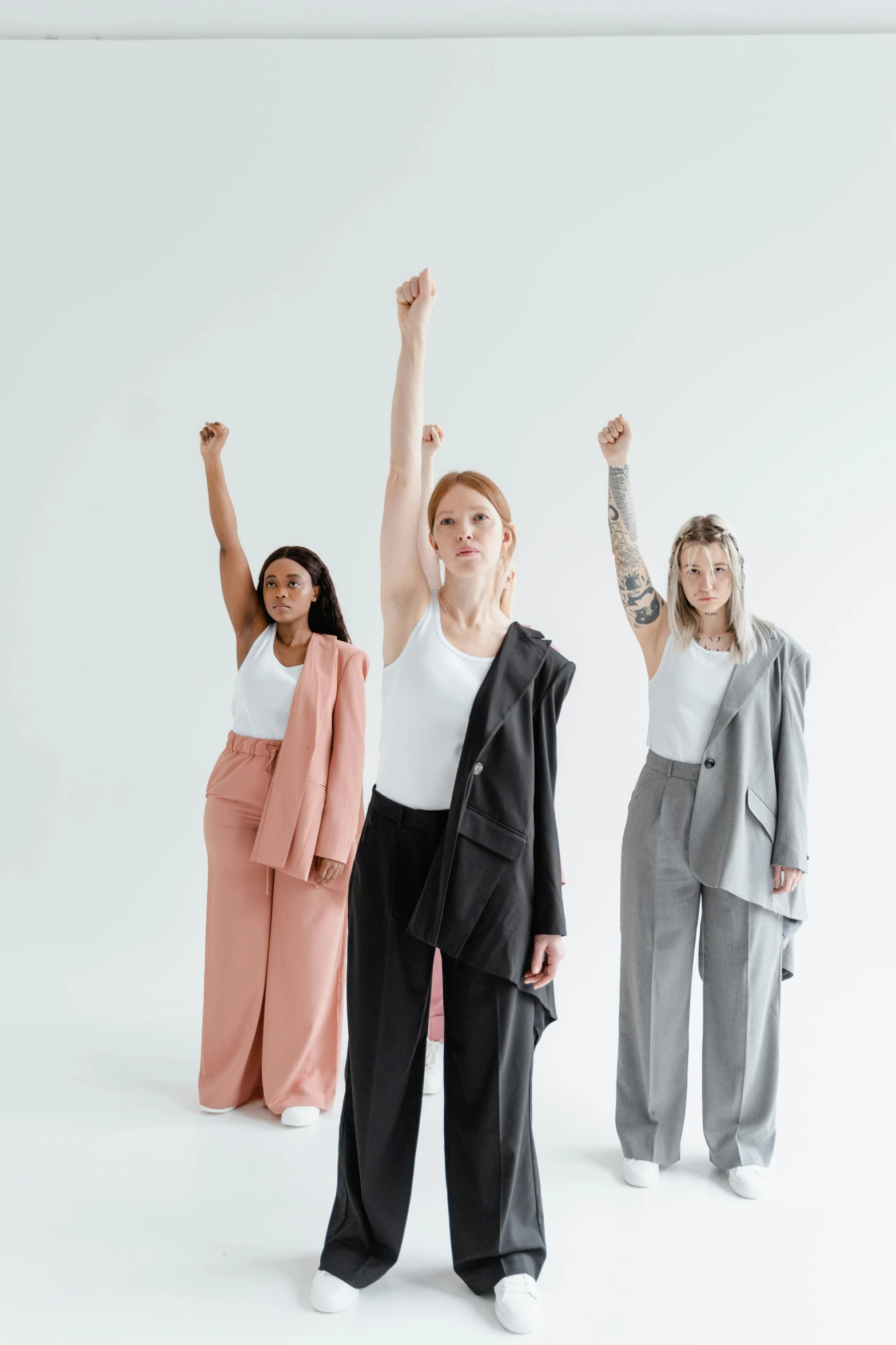 a group of women standing in front of a white wall, trending on pexels, feminist art, three piece suit, one fist raised high in triumph, realistic human movement, on a white table