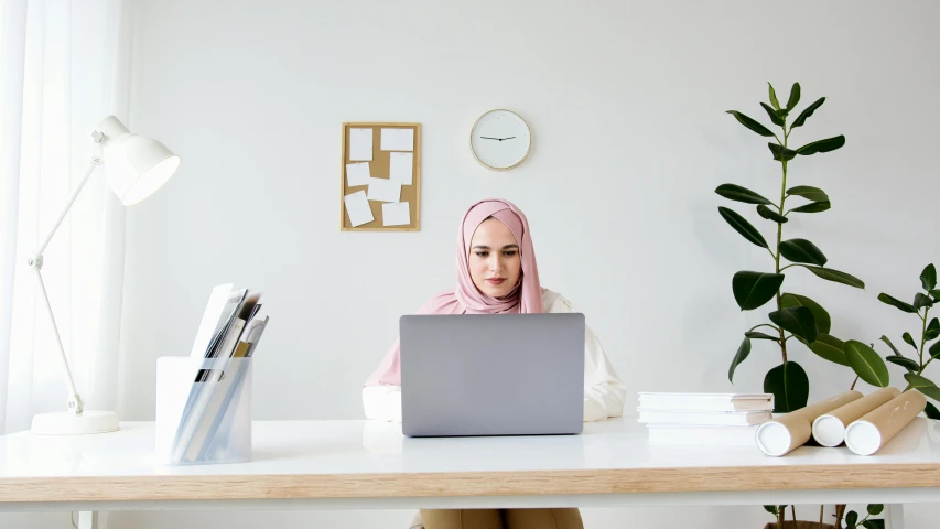 a woman sitting at a desk using a laptop computer, a picture, inspired by Maryam Hashemi, trending on pexels, hurufiyya, white and pink cloth, looking content, slightly minimal, islamic