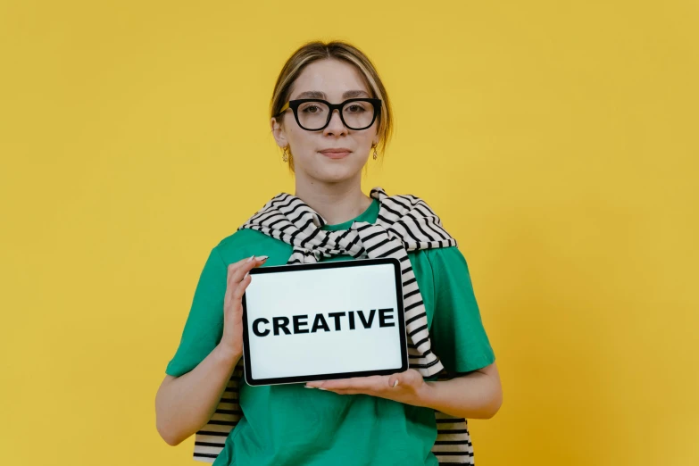 a woman holding a sign that says creative, by Julia Pishtar, trending on pexels, interactive art, in square-rimmed glasses, on a yellow canva, costume desig, 8 k what