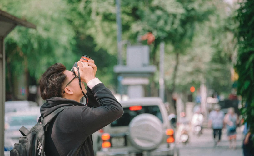 a man taking a picture of a busy street, pexels contest winner, goggles on forehead, looking to the sky, asian male, wearing black frame glasses