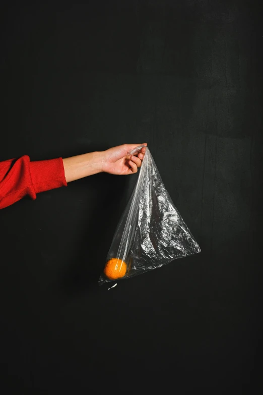 a person holding an orange in a plastic bag, on a black wall, no - text no - logo, 9