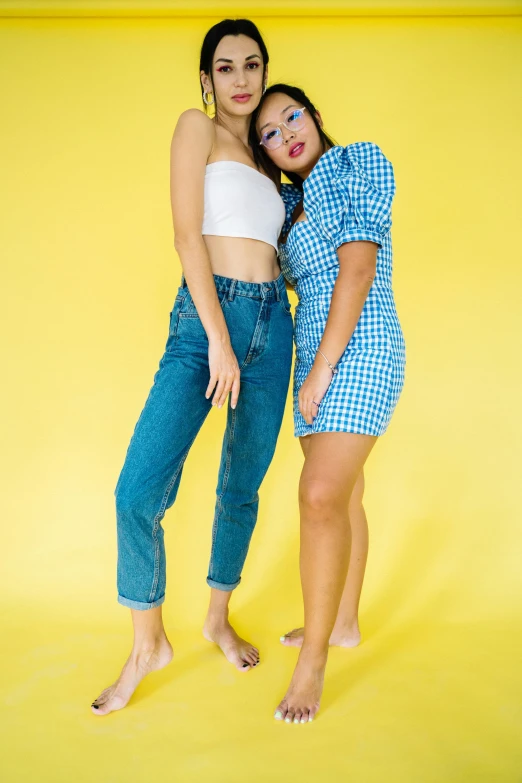 two women standing next to each other on a yellow background, by Juan O'Gorman, trending on pexels, daisy dukes, blue checkerboard dress, ( ( ( wearing jeans ) ) ), studio shoot