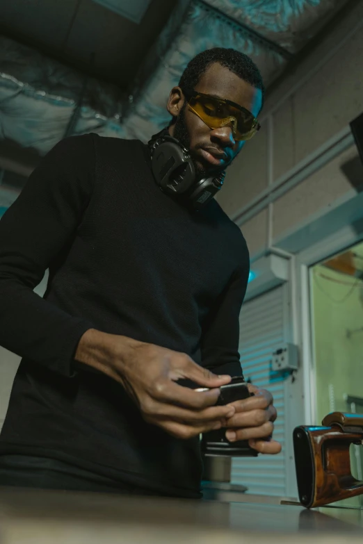 a man standing next to a table holding a cell phone, inspired by Terrell James, trending on pexels, afrofuturism, gaming headset, wearing a black sweater, in a studio, wearing black rimmed glasses