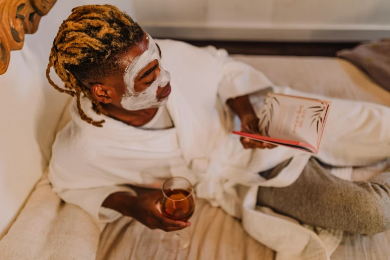 a woman sitting on a bed holding a glass of wine, by Julia Pishtar, pexels contest winner, african mask, skincare, male with halo, reading for a party