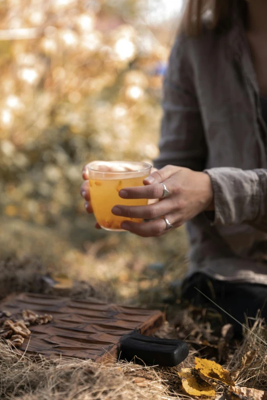 a woman sitting on the ground holding a cup of tea, by Jessie Algie, trending on unsplash, renaissance, holding a large glass of beer, cider - man, forest picnic, hand on table