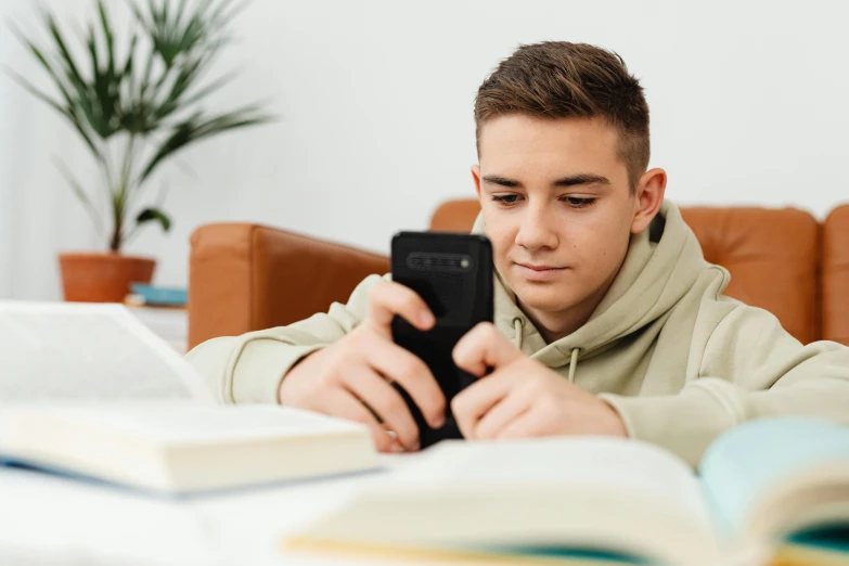 a man sitting at a table looking at a cell phone, textbooks and books, rated t for teen, boy with neutral face, instagram post