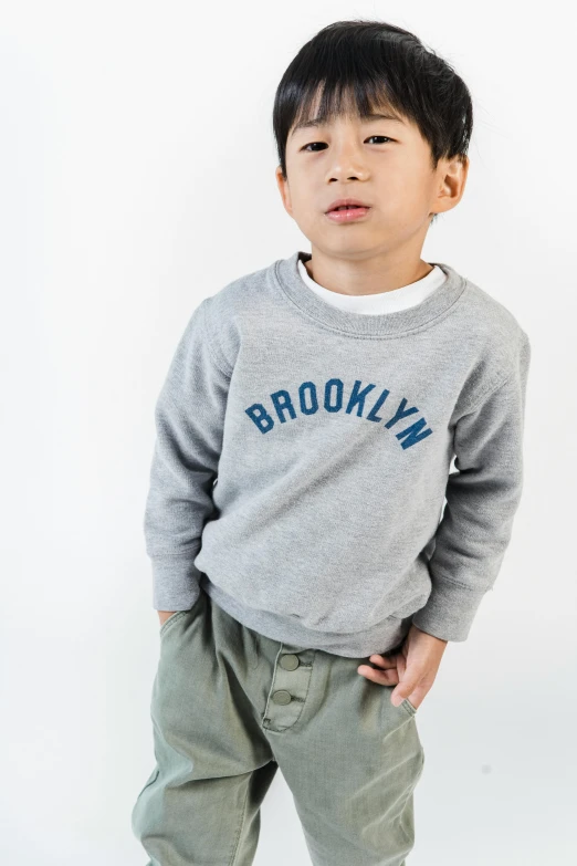 a young boy standing with his hands in his pockets, a colorized photo, trending on unsplash, brooklyn background, trendy typography, grey sweater, toddler