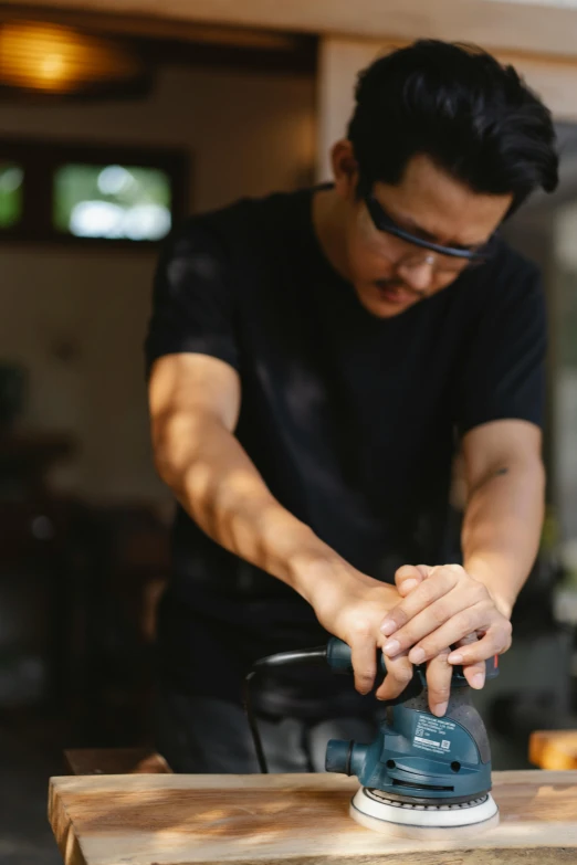 a man sanding a piece of wood with a sander, inspired by Hiroshi Honda, unsplash, wearing black frame glasses, picture of a male biker, partially cupping her hands, asian male