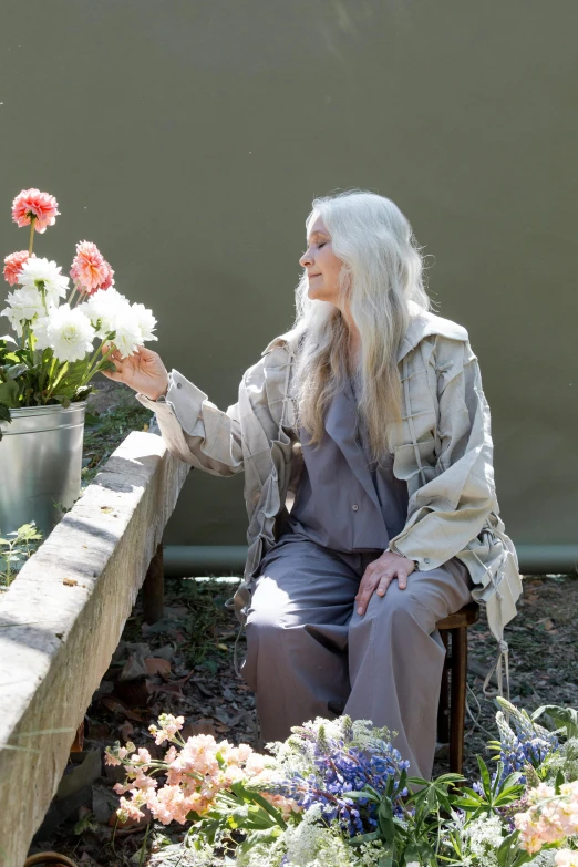 a woman sitting on a bench in a flower garden, a portrait, inspired by Grethe Jürgens, unsplash, photorealism, light gray long hair, film still promotional image, carrying flowers, aging