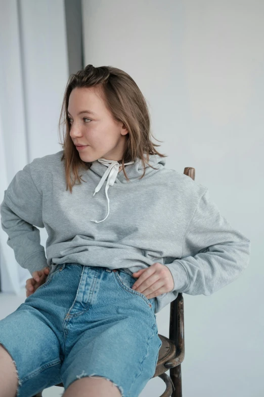 a woman sitting on top of a wooden chair, inspired by Louisa Matthíasdóttir, trending on pexels, happening, grey hoodie, denim, thick collar, teenager