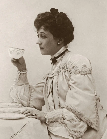 a woman sitting at a table drinking from a cup, a black and white photo, by Rose Maynard Barton, art nouveau, wearing 1890s era clothes, promo image, with shoulder pads, auguste toulmouche
