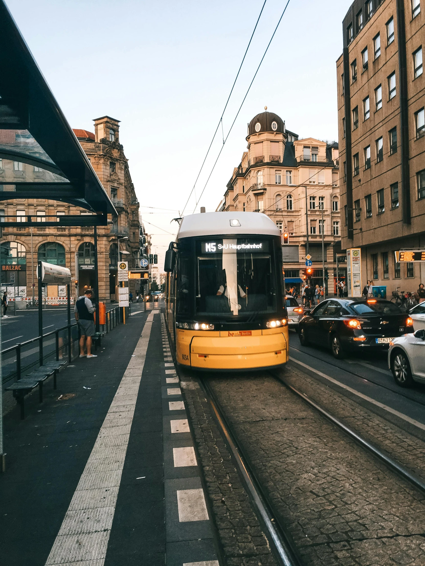 a yellow train traveling down a street next to tall buildings, by Matija Jama, pexels contest winner, tram, late summer evening, square, 🚿🗝📝