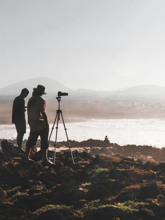 a group of people standing on top of a rocky beach, a picture, pexels contest winner, happening, two still figures facing camera, video footage, silhouettes in field behind, tripod