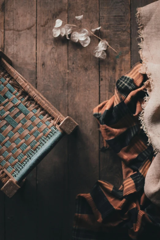 a blanket sitting on top of a wooden floor next to a basket, a still life, by Jessie Algie, trending on unsplash, lumberjack, woven armour, brown and cyan color scheme, rags