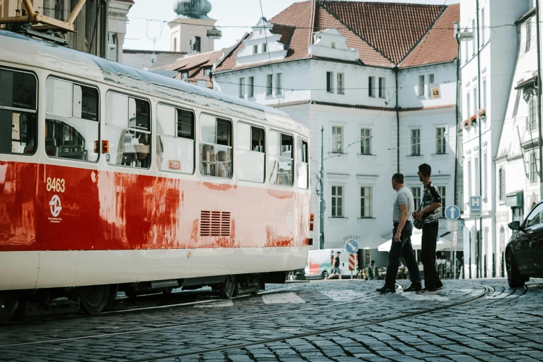 a couple of people standing next to a red and white train, by Emma Andijewska, pexels contest winner, viennese actionism, trams ) ) ), square, cobblestone streets, summer light