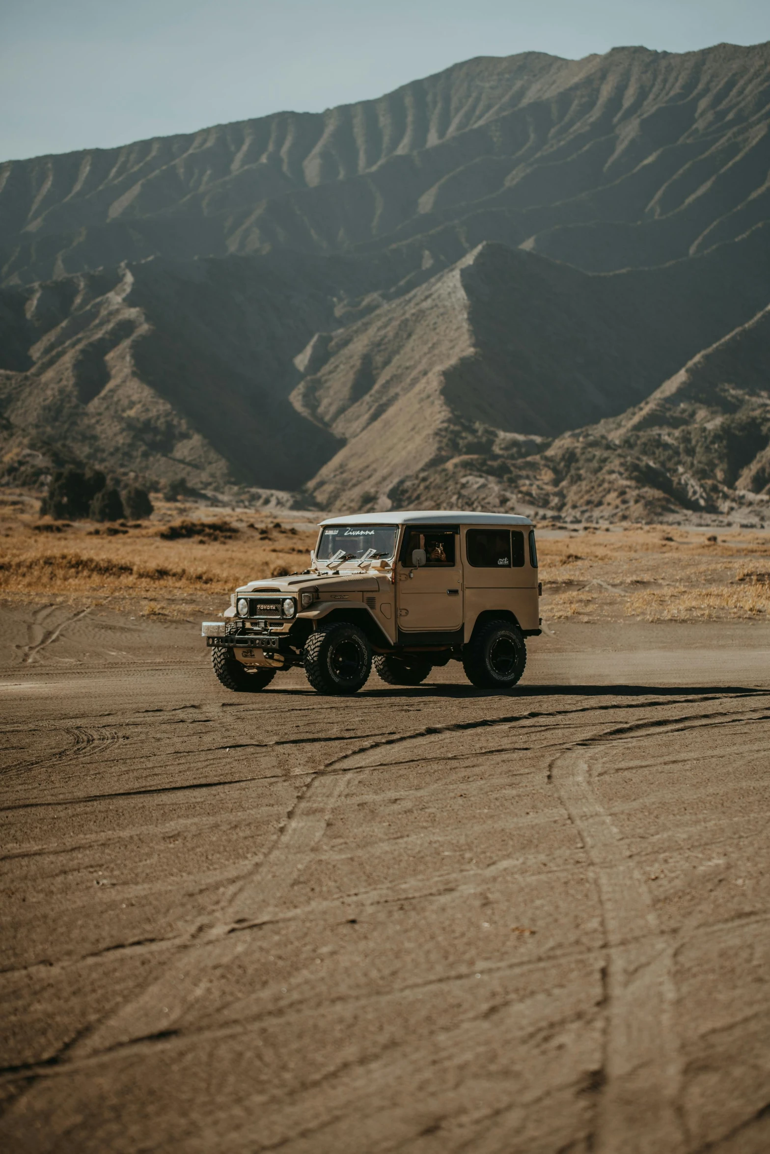 a jeep driving down a dirt road with mountains in the background, unsplash contest winner, minimalism, brown and cream color scheme, square, indonesia, classic cars