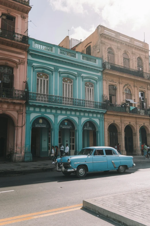 a blue car driving down a street next to tall buildings, a colorized photo, trending on unsplash, baroque, cuban revolution, ornate palace made of green, faded worn, square