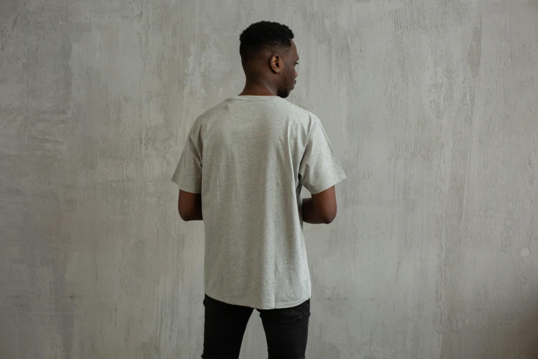 a man standing in front of a concrete wall, inspired by Paul Georges, visual art, printed on a cream linen t-shirt, view from back, dressed in a gray, looking left