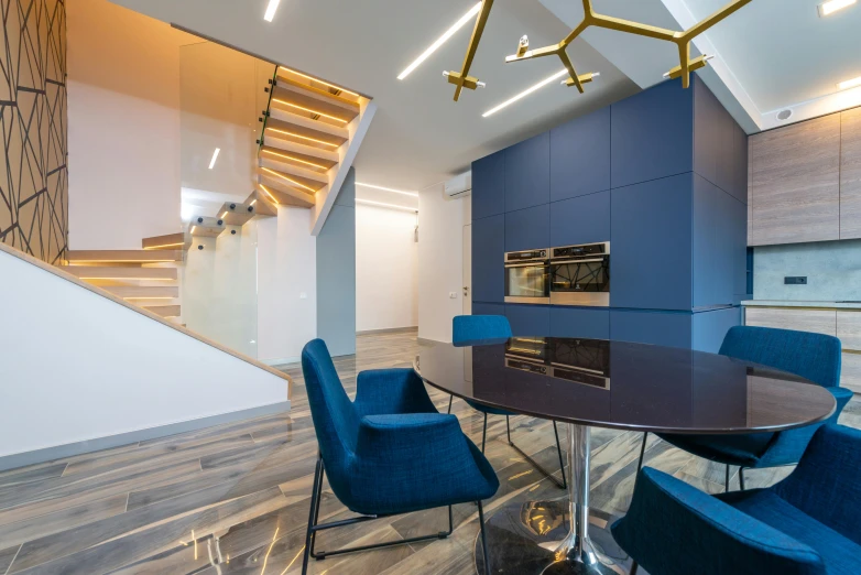 a dining room with a round table and blue chairs, by Adam Marczyński, pexels contest winner, light and space, stairs to an upper floor, modern office, accented in bright metallic gold, angular asymmetrical design