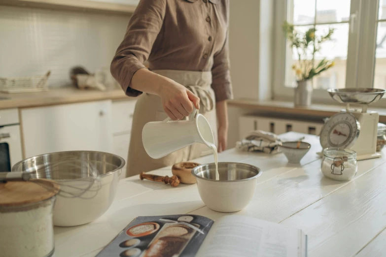 a woman pouring milk into a bowl in a kitchen, inspired by Wilhelm Hammershøi, trending on pexels, white clay, stainless steel, pastry, chef table