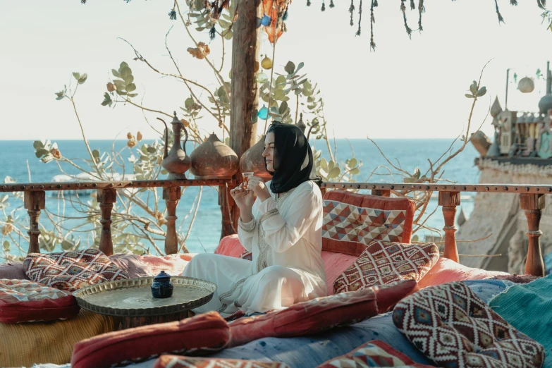 a woman sitting on top of a bed next to the ocean, inspired by Riad Beyrouti, pexels contest winner, arabesque, celebration of coffee products, sitting in a cafe, sheikh, cottagecore hippie