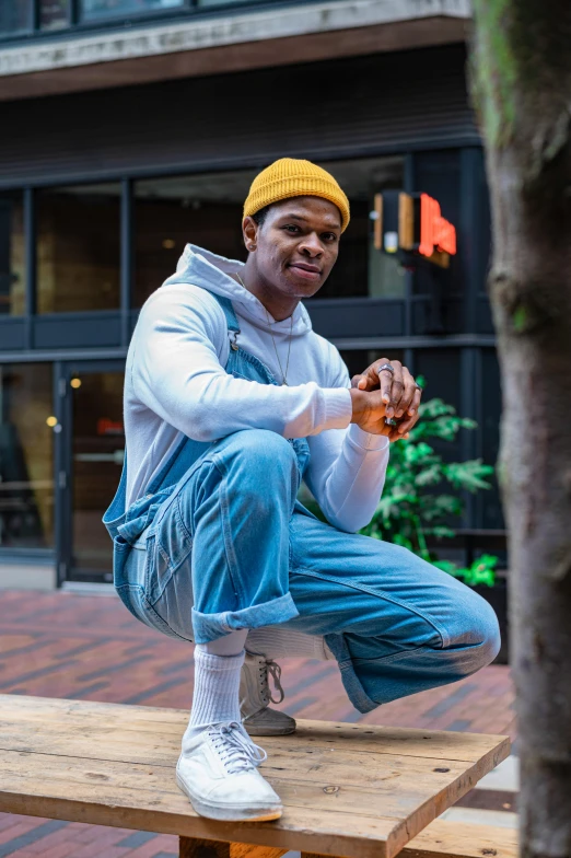 a man sitting on top of a wooden bench, blue overalls, wearing teal beanie, david uzochukwu, doing a sassy pose