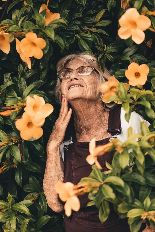 an older woman standing in front of a bush of flowers, by Elsie Few, pexels contest winner, wrinkled skin, looking upward, instagram photo, high angle shot