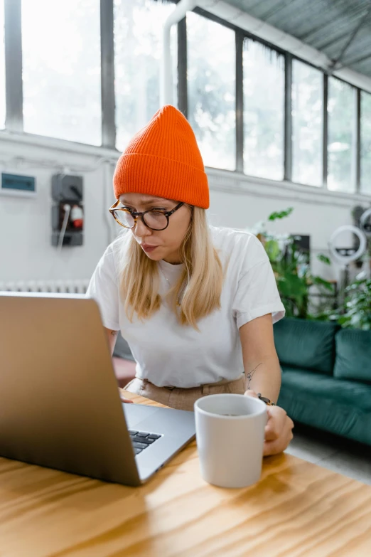 a woman sitting at a table working on a laptop, trending on pexels, beanie, white and orange, nerdy appearance, gif