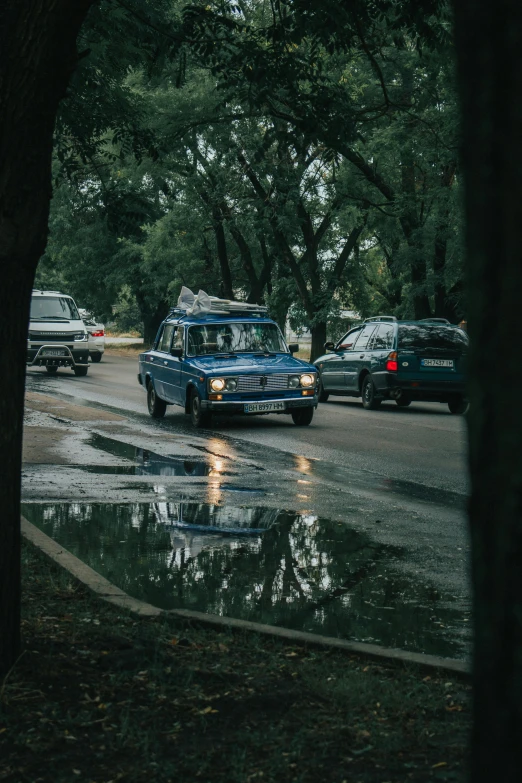 a group of cars driving down a street next to trees, a picture, unsplash contest winner, realism, puddle of water, russian lada car, in a suburb, analog photo